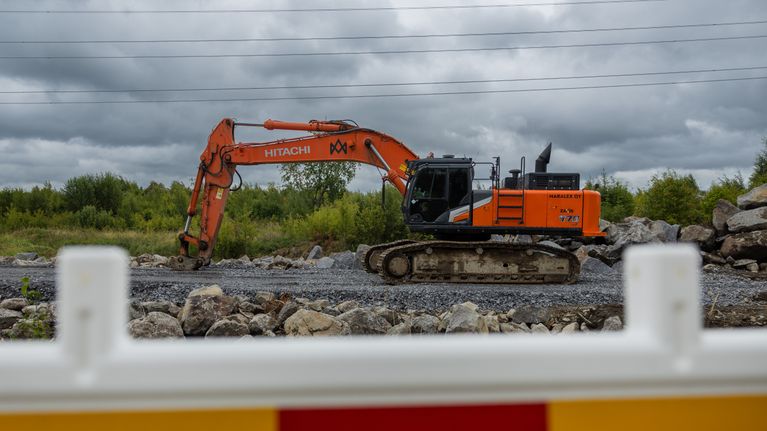 En orange grävmaskin arbetar på en grusväg, stenar och elledningar syns i bakgrunden.