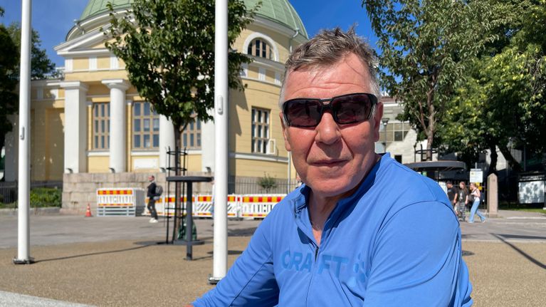 Mies katsoo kameraa kohti sinisessä asussa istuessaan Turun Kauppatorin penkeillä.