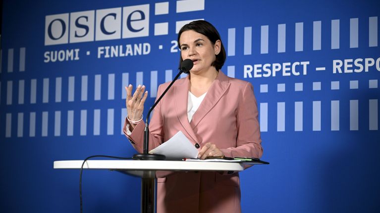 Nainen seisoo puhujapöydän ääressä mikrofonin edessä, taustalla OSCE-tapahtuman seinä.