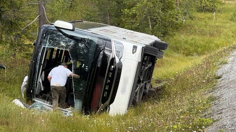 Isompi valkoinen linja-auto on kaatunut kyljelleen tien penkalle, ja mies seisoo bussin oven kohdalla.
