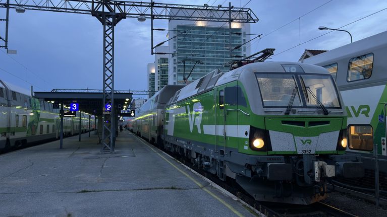 Vihreävalkoinen VR-juna seisoo asemalaiturilla, taustalla korkea rakennus ja ratapihan opasteissa näkyy tekstiä.