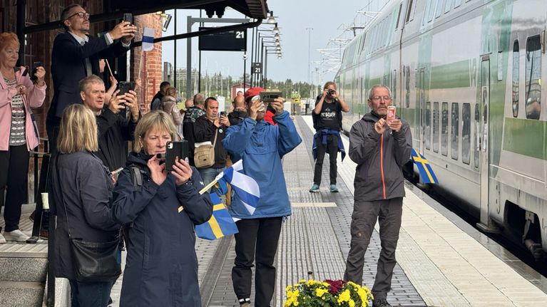 Ihmisiä seisoo rautatieasemalla ottamassa kuvia saapuvasta vihervalkoisesta junasta, esillä Suomen ja Ruotsin lippuja.