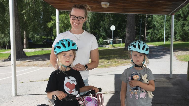 Aikuinen ja kaksi lasta seisovat ulkona pyörätelineen vieressä, lapsilla pyöräilykypärät ja toisella polkupyörä.