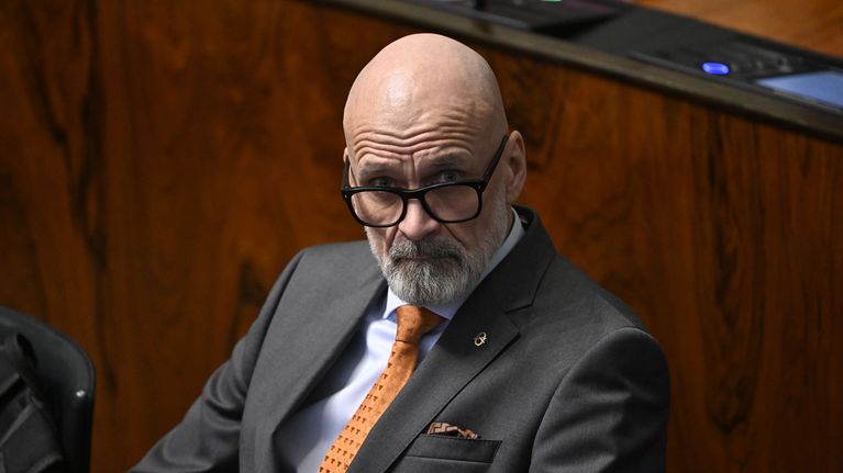 Pukuun ja oranssiin solmioon pukeutunut silmälasipäinen mies istuu puupintaisessa salissa pöydän ääressä.