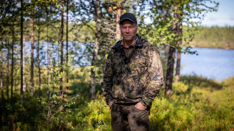 Mies seisoo metsäisessä maisemassa järven rannalla, taustalla puita ja vettä.