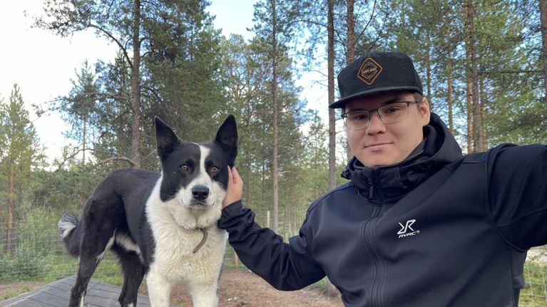 Riku Rasmus seisoo ulkona metsämaisemassa käsi koiran pään päällä, taustalla aitauksia ja puita.