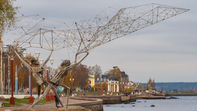 Rantapromenadilla sijaitseva metallista valmistettu suuri kalastajapatsas, taustalla vesistöä ja rakennuksia.