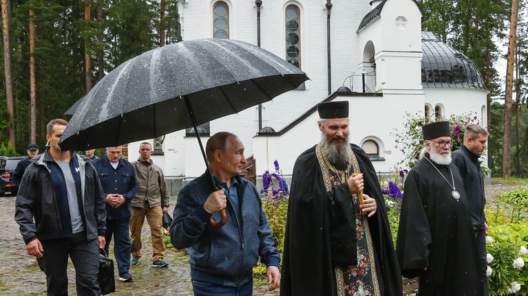 Useita ihmisiä kävelee sateessa luostarin pihalla, taustalla valkoinen kirkko ja puita.