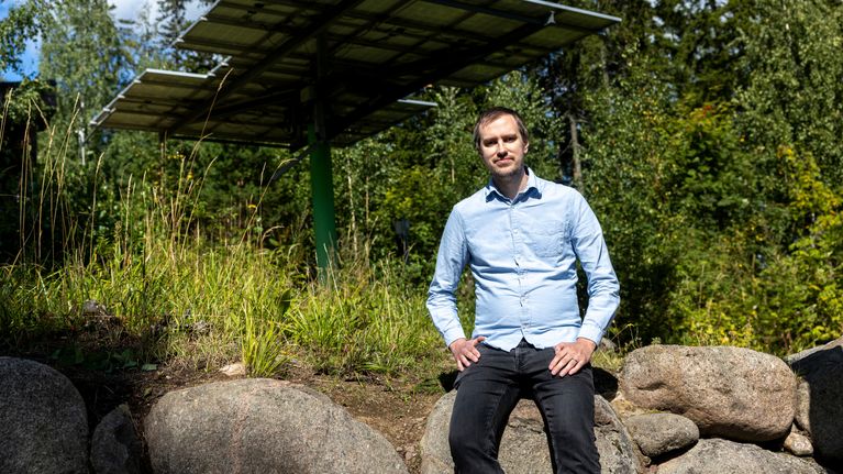 Mies seisoo kivillä aurinkopaneelin edessä metsäisessä ympäristössä, taustalla puita ja pilvinen taivas.