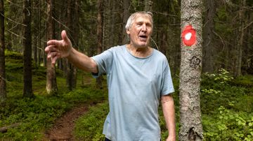 Vanha mies viittoo kädellä metsässä puun vieressä, jossa on punainen reittimerkki.