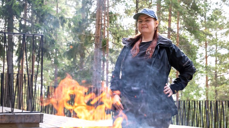 Nainen seisoo laavulla ja hänen edessään on nuotion liekkejä ja savua.