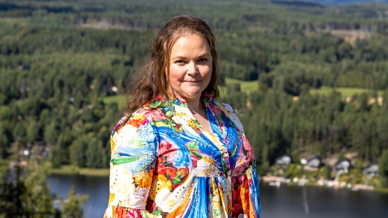 Nainen seisoo värikkäässä mekossa korkean mäen päällä taustallaan järvi ja metsikkö.