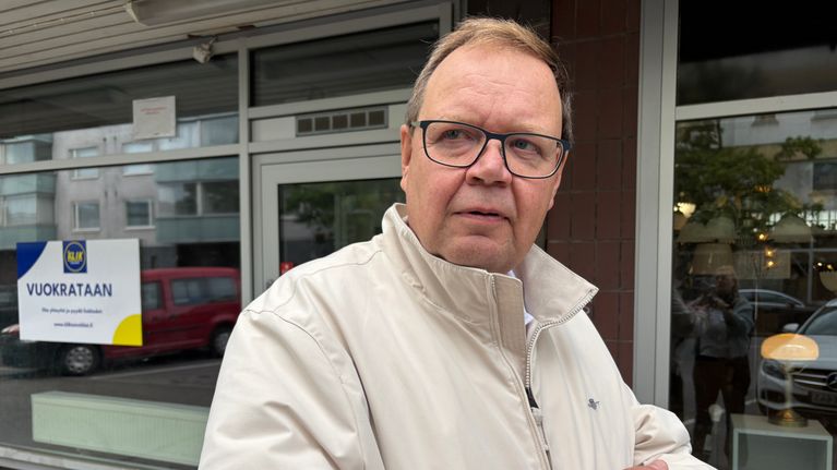 Mies seisoo kadulla rakennuksen edessä, taustalla ikkunassa näkyy "VUOKRATAAN" -kyltti.