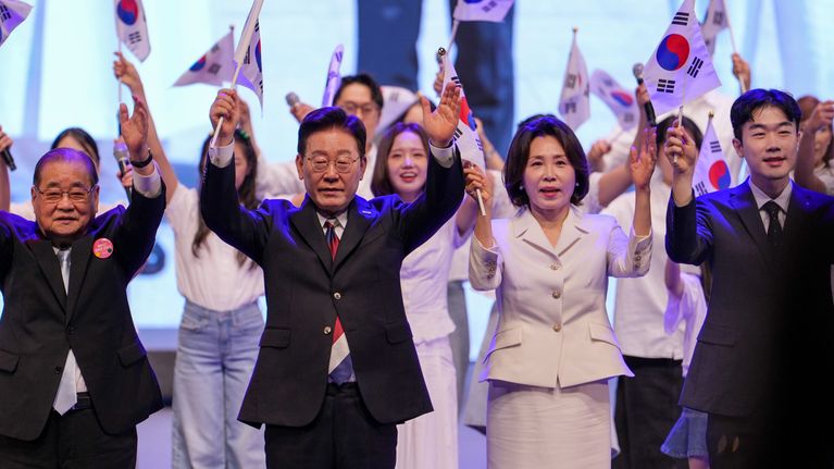 Useita ihmisiä seisoo rivissä ja heiluttaa Etelä-Korean lippuja; taustalla ryhmä nuoria valkoisissa vaatteissa.