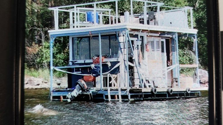 Kelluva, kaksikerroksinen saunalautta järvellä, yksi henkilö työskentelee moottorin parissa.