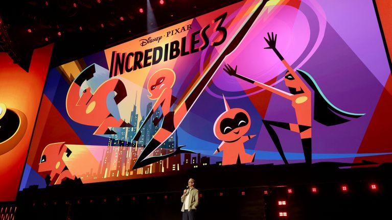 Henkilö seisoo lavalla suuren valotaulun edessä, jossa lukee "Disney Pixar INCREDIBLES 3" ja näkyy piirroshahmoja.