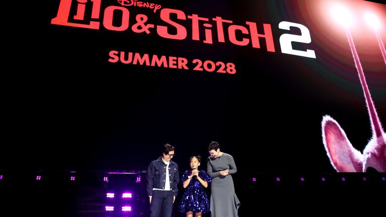 Kolme ihmistä seisoo lavalla ison näytön edessä, jossa lukee ”Lilo & Stitch 2 SUMMER 2028”.