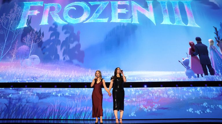 Kaksi naista seisoo lavalla ja puhuu mikrofoneihin, taustalla suuri "Disney Frozen III" -teksti ja animaatiokuva.