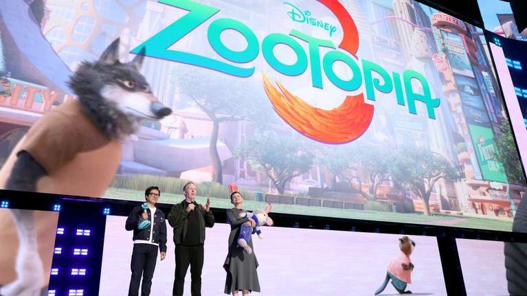 Kolme ihmistä ja eläinhahmo lavalla suuren Zootopia 2 -elokuvajulisteen edessä.