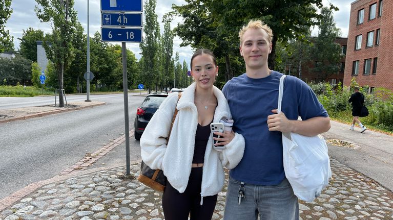 Kaksi nuorta aikuista seisoo kadun varrella pysäköintimerkin edessä; toisella on juomamuki kädessä.