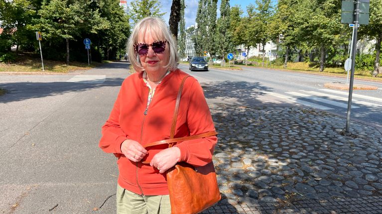 Nainen seisoo aurinkoisella kadulla puiden ja suojatien läheisyydessä, yllään oranssi takki ja ruskea laukku.
