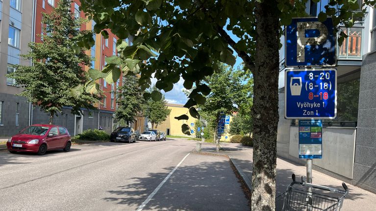 Kadun varrella pysäköityjä autoja, pysäköintikyltti, tyhjä ostoskärry ja tien vieressä puita.
