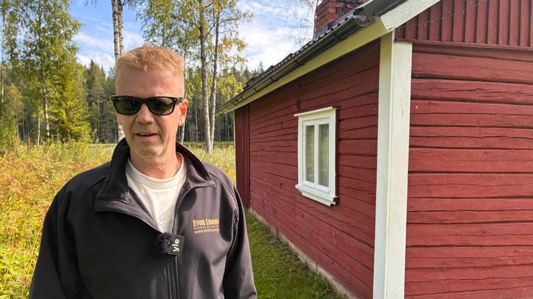Mies seisoo aurinkolasit päässä punaisen saunarakennuksen edessä, takissa lukee Evon luonto.