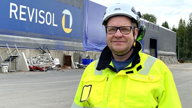 Suojakypärään ja työvaatteisiin pukeutunut, silmälasipäinen mies seisoo suuren pressuhallin vieressä.