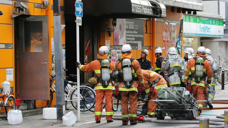 Seitsemän palomiestä seisoo rakennuksen sisäänkäynnin edustalla varusteissaan, poliiseja taustalla.