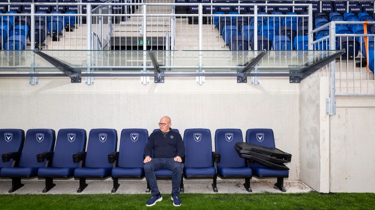 Mies istuu yksin jalkapallostadionin vaihtopenkillä, taustalla tyhjät katsomorivit.