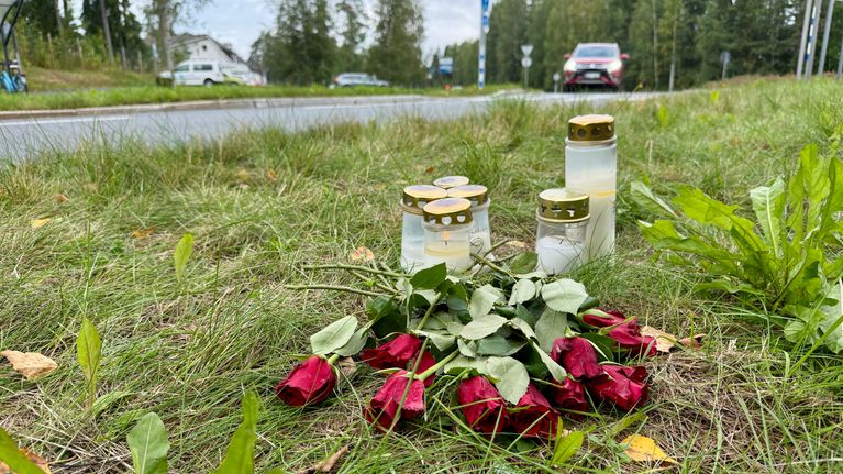 Kynttilöitä ja punaisia ruusuja tienvarren ruohikolla, taustalla autotie ja autoja.