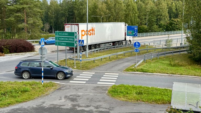 Henkilöauto ja Posti-rekka liikenneympyrän lähellä, etualalla ramppia ylittävä suojatie, liikennemerkeissä tekstit Helsinki, Hämeenlinna ja Turku.