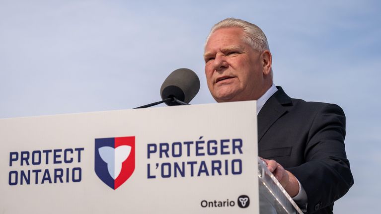 Harmaahiuksinen pukuun pukeutunut mies puhujapöntössä, jossa lukee "Protect Ontario"