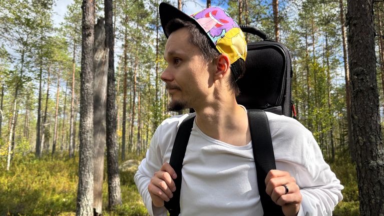 Mies seisoo metsässä, päässään värikäs lippalakki ja selässään reppu. Taustalla näkyy puita ja vihreää maastoa.