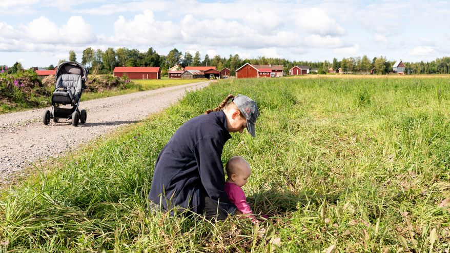 Aikuinen ja lapsi istuvat nurmikolla maaseudulla, taustalla maatilan rakennuksia ja hiekkatie, jonka varrella on lastenvaunut.