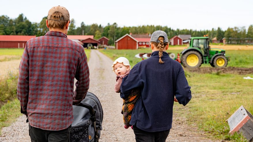 Kaksi aikuista ja pieni lapsi kulkevat hiekkatietä maatilalla, taustalla näkyy traktori ja punaisia rakennuksia.