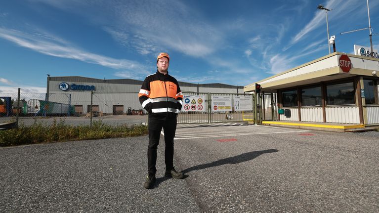 Škoda Transtech Otanmäen tehtaan pääluottamusmies Mikko Komulainen oranssi-mustassa työvaatteessa seisoo tehdasalueen portin edessä, taustalla tehdas.