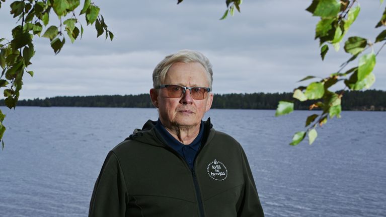 Mies seisoo hiekkarannalla järven rannalla, taustalla vettä ja pilvinen taivas, edustalla koivunoksia.