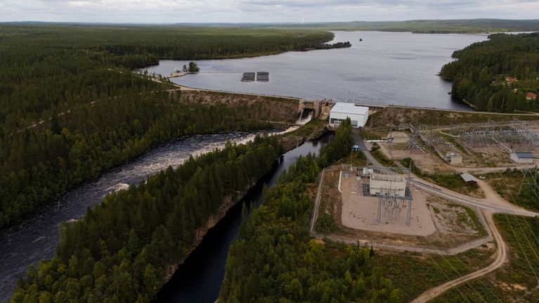 Ilmakuva vesivoimalaitoksesta joen ja järven rannalla, ympärillä metsää ja sähköaseman rakenteita.