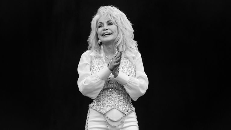 Dolly Parton seisoo lavalla ja katsoo eteenpäin, pukeutuneena valkoisiin koristeellisiin vaatteisiin.