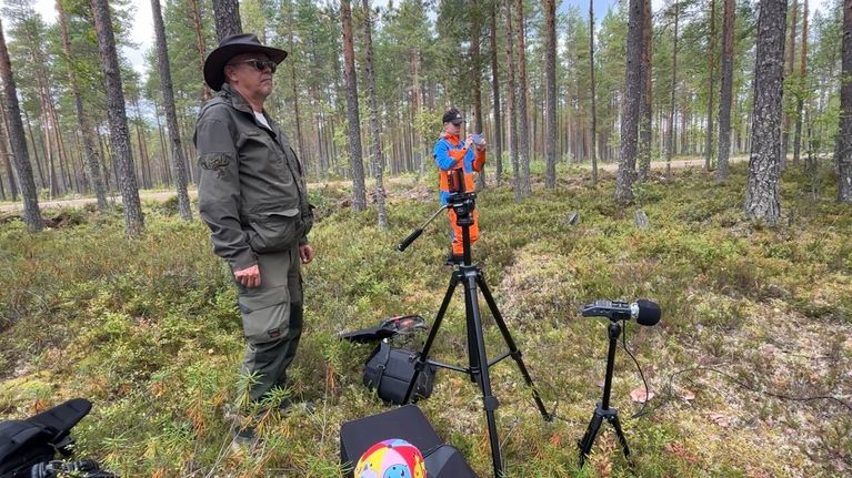 Kaksi henkilöä seisoo metsässä kamerajalustojen ja äänityslaitteiden vieressä, taustalla puita ja sammalta.