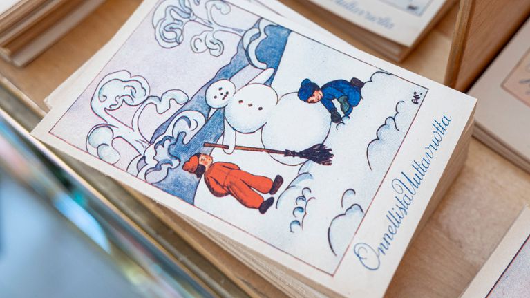 Postikortteja myyntitelineessä, etualalla kortti, jossa kaksi lasta tekee lumiukkoa ja teksti "Onnellista uutta vuotta".