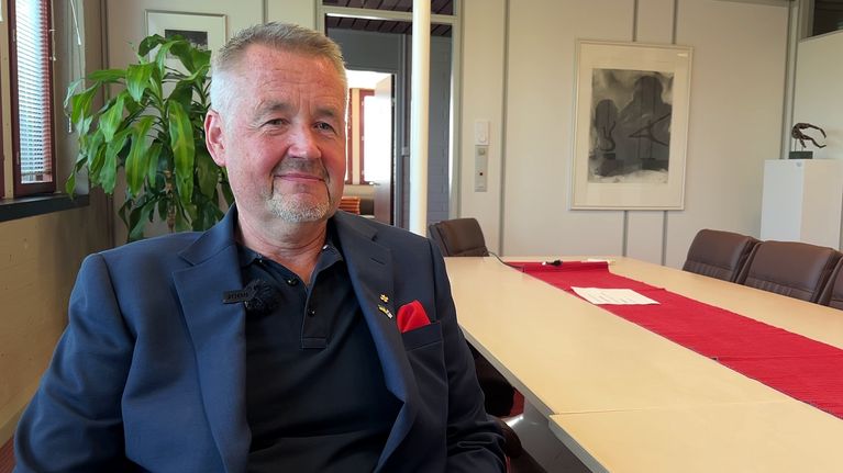 Mies tumma puku yllään istuu neuvotteluhuoneessa pitkän pöydän päässä. Taustalla taulu ja kasvi.
