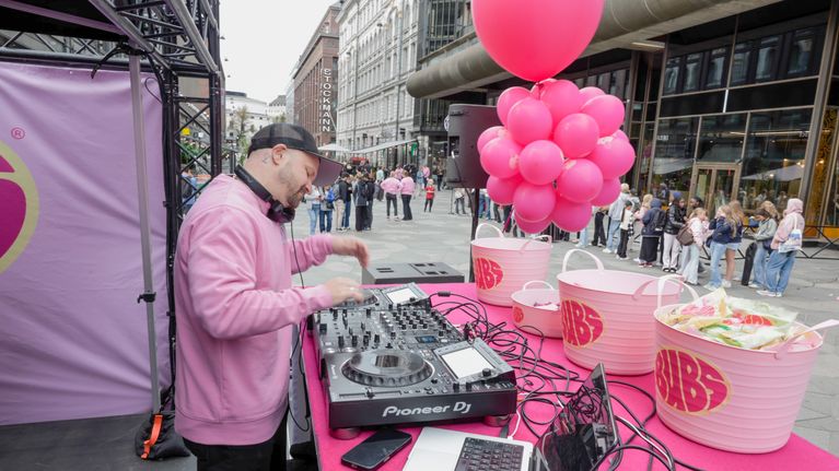 DJ soittaa musiikkia ulkoilmatapahtumassa vaaleanpunaisen pöydän ääressä, ympärillä ilmapalloja ja ihmisjoukko.