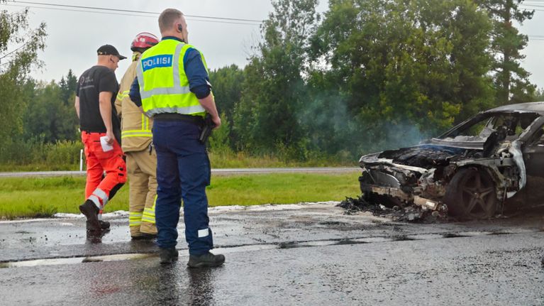 Poliisi, palomies ja pelastaja tutkivat tien varrella palanutta autoa, taustalla näkyy metsää.
