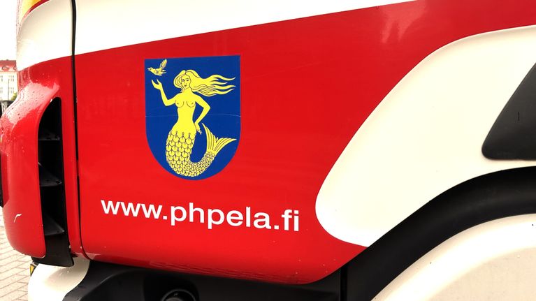 Lähikuvassa on paloauton etuovi, jossa on punaista, valkoista sekä sinikeltainen vaakunta. Tekstinä lukee www.phpela.fi.