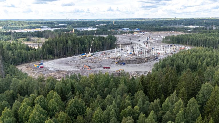 Laaja rakennustyömaa metsän keskellä, useita nostureita ja työajoneuvoja avoimella alueella.