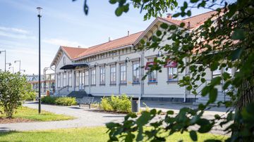 Vaalea punakattoinen puurakennus, pihalla nurmikkoa, puita ja istutuksia.
