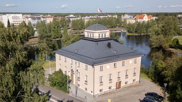 Ilmakuvassa vaalea rakennus, jossa musta katto ja katolla liehuu lippu. Taustalla joki ja kaupunkia.