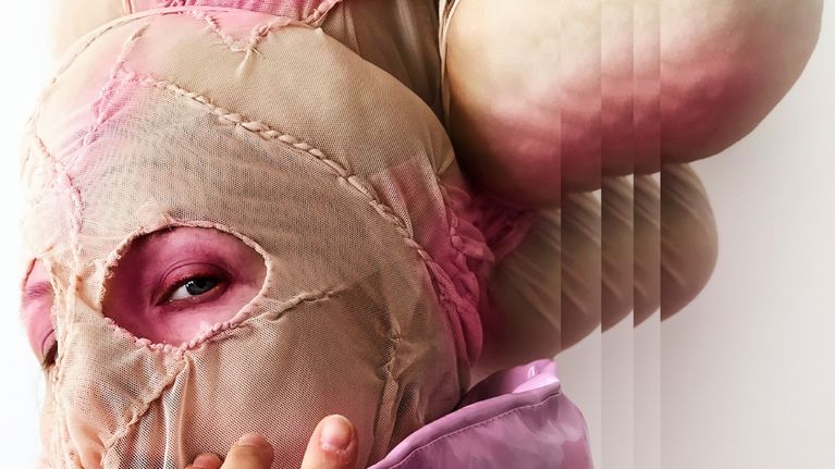 Henkilö pukeutuneena vaaleanpunaiseen takkiin ja vaaleaan, ommeltuun naamioasuun; kasvot osittain peitetty.